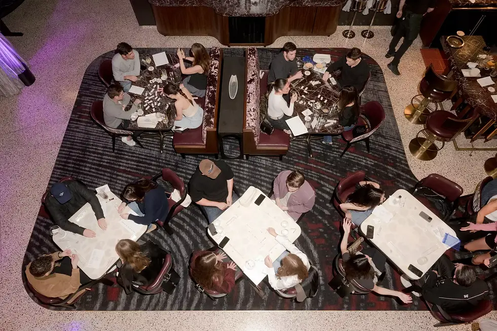 People seated at tables in a restaurant, engaged in conversation, on a patterned carpeted floor. Background shows bar area and dim lighting.