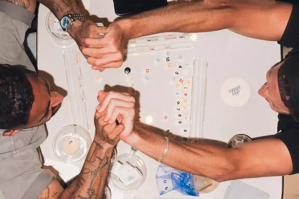 Two men arm wrestling over a table with game tiles scattered. One has tattoos and a watch. Text "runni klub" on a coaster. Bright setting.