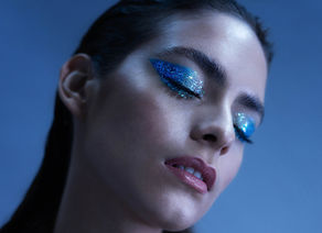 Woman with eyes closed, wearing glittery blue eyeshadow in a contemplative pose. Background is a soft blue, wearing a maroon jacket.