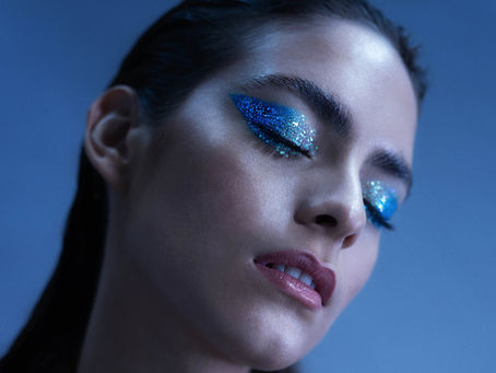 Woman with eyes closed, wearing glittery blue eyeshadow in a contemplative pose. Background is a soft blue, wearing a maroon jacket.