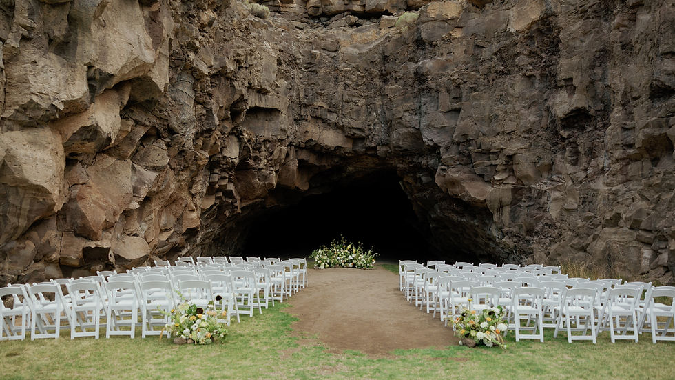 Chairs and flowers set up for a wedding in front of a large cave entrance. Dreamy Green and White Flowers Inspirations with brown rock walls and grassy area create a natural setting.