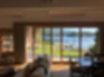 Photograph taken from the interior of a house overlooking a lake and a green garden in the front.