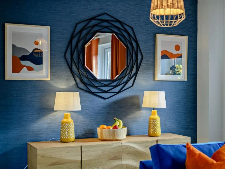 A blue feature wall with a black mirror, sideboard and yellow lamps