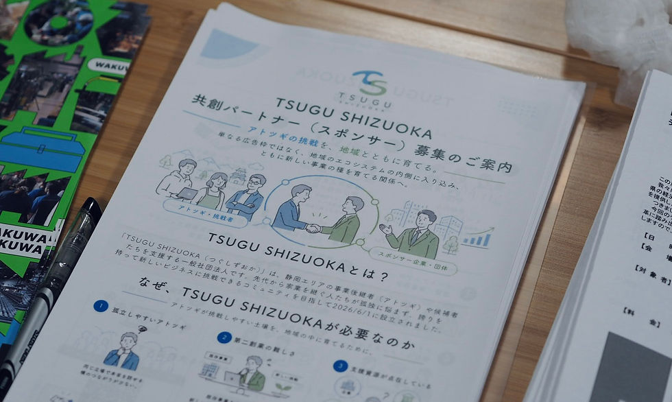 木のテーブルに置かれた「TSUGU SHIZUOKA」スポンサー募集案内の冊子。握手や人物イラストがあり、左に緑のパンフとペンが見える。