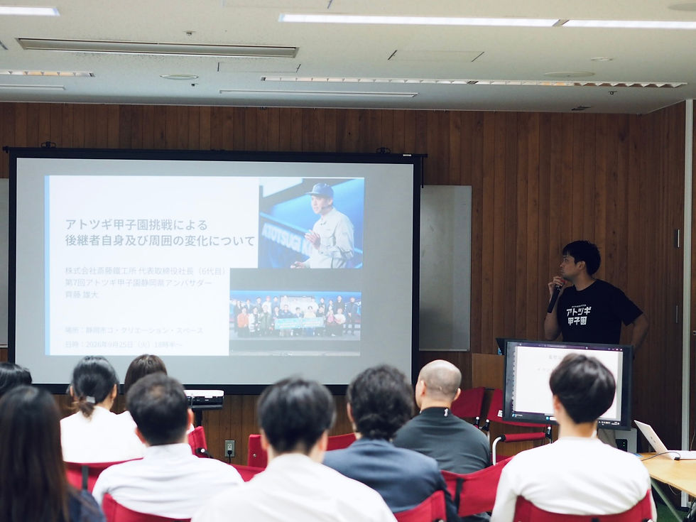 木目の会議室で、黒Tシャツの男性がマイクで発表し、前方のスクリーンに「アトツギ甲子園」挑戦のスライドが映っている。