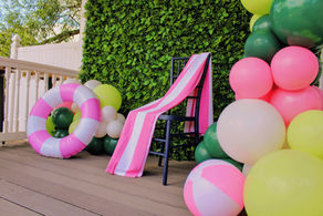 Pink-and-white chair and pool float among pastel balloons on a deck by a green plant wall, festive outdoor party setup