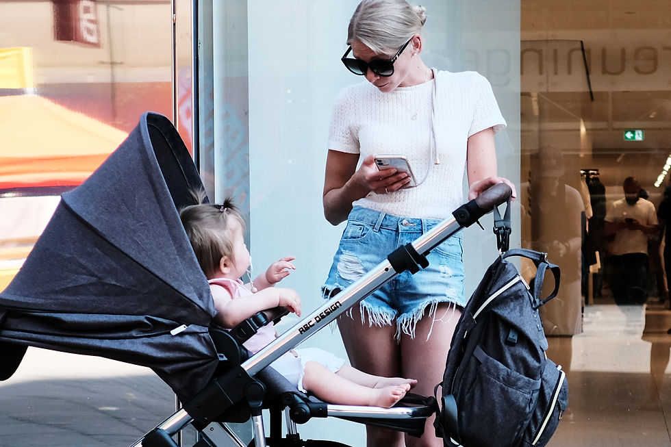Frau in weißem Oberteil schaut aufs Handy neben Kinderwagen mit Baby. Jeansshorts, Sonnenbrille. Einkaufsumgebung, entspanntes Szenario.