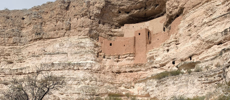 A five-story cliff dwelling built into a limestone cliff, Montezuma Castle stands above the desert floor with small, square windows and adobe-colored walls. Surrounded by sparse vegetation and set against a bright blue Arizona sky.