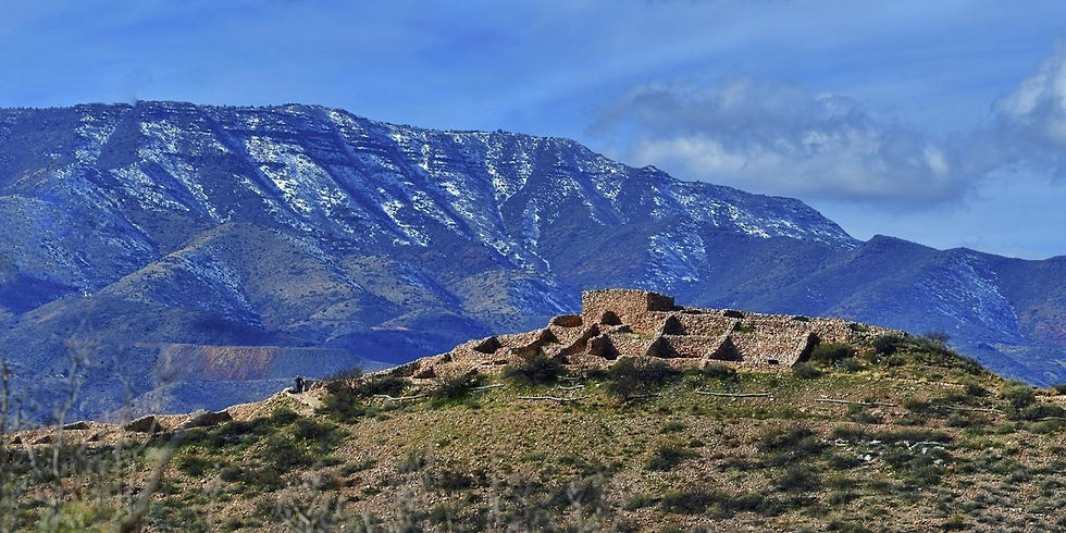 The stone ruins of Tuzigoot National Monument stretch across a desert hilltop, with snow-capped mountains rising in the distance. The ancient pueblo’s tan masonry contrasts with the crisp white peaks and clear sky, highlighting the site’s striking setting.