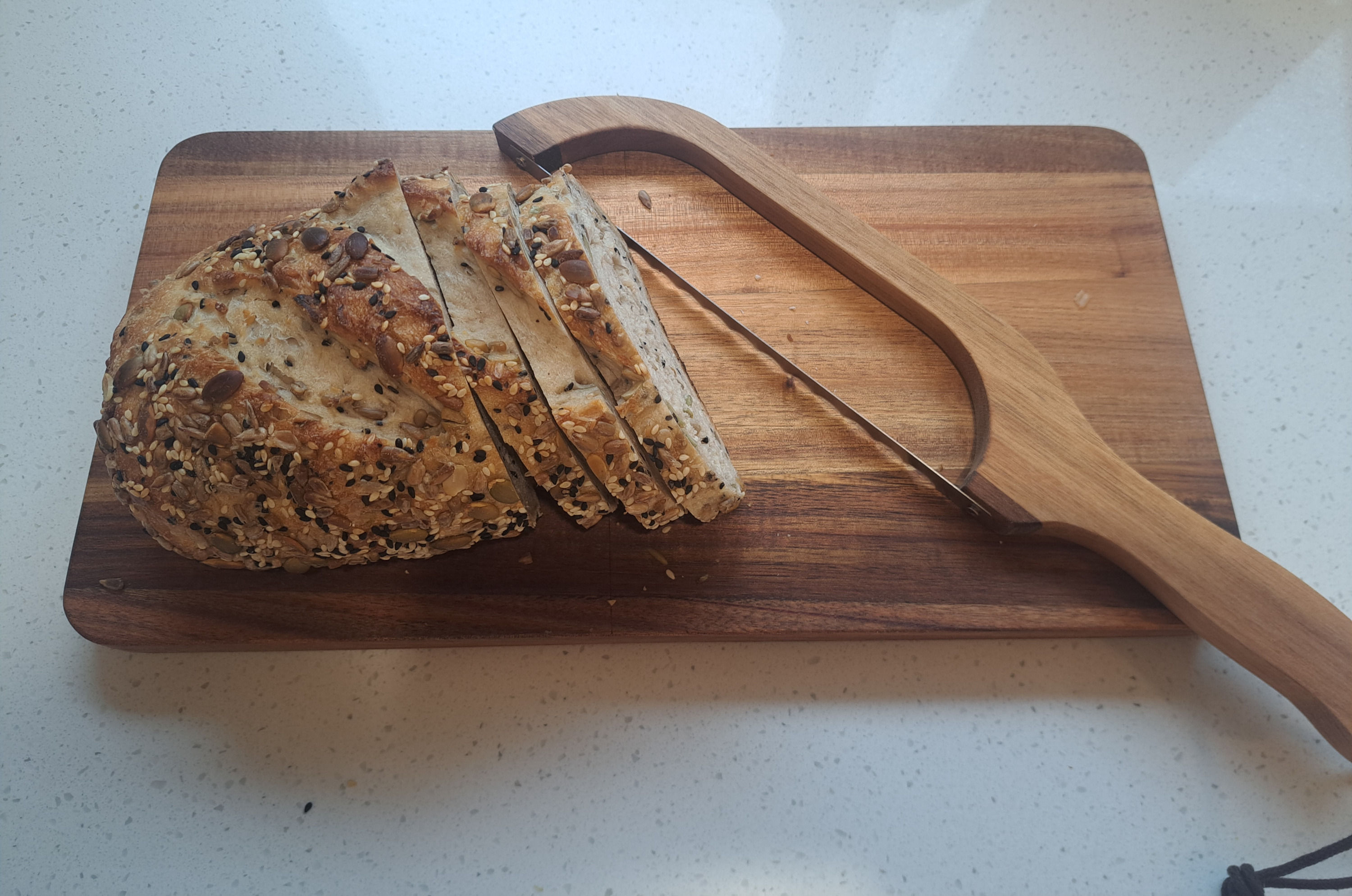 Seeded sourdough loaf and slices on an acacia cutting board with a wooden bow bread knife.