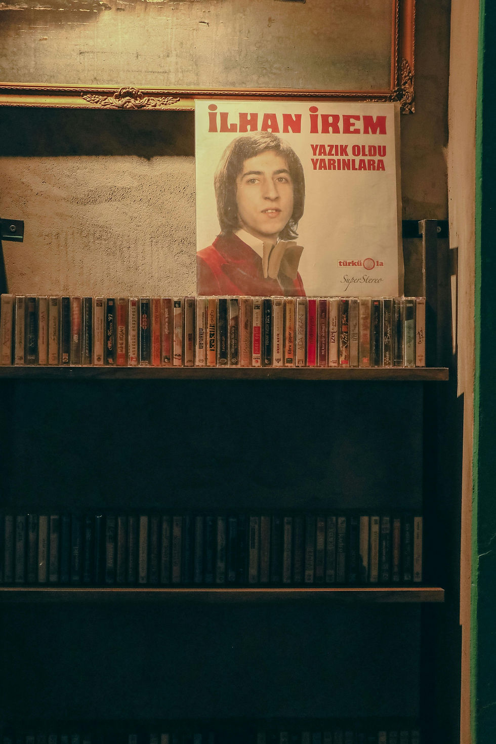 A vintage music collection displayed in a cozy setting, featuring a retro poster of a classic album by İlhan İrem above a shelf lined with cassette tapes.