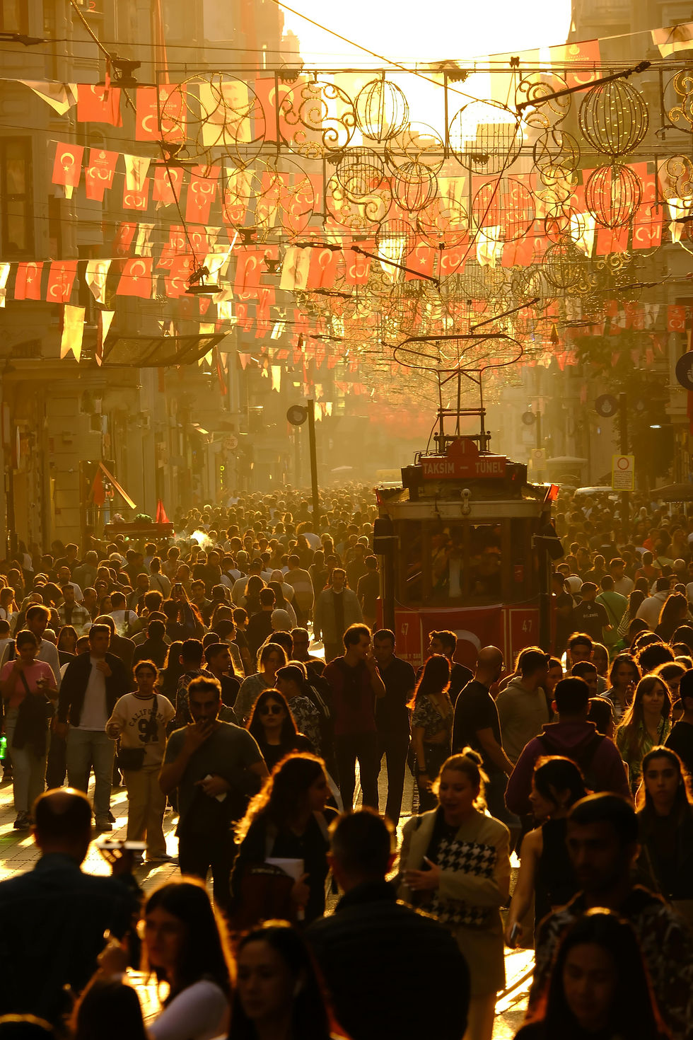 A bustling Istiklal Street in Istanbul, alive with people under the glow of a setting sun, as a historic tram makes its way through the vibrant crowd adorned with Turkish flags.