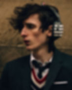 Modelo masculino de perfil usando los audífonos de diadema Thom Browne x Beats con patrón de rayas blancas y traje de sastre con suéter de cuello V escolar