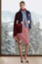 A full-length fashion editorial shot of a model standing on a light wood floor in a room with a large window. Through the window, a panoramic view of snowy mountains and pine forests is visible. The model wears a carefully curated, layered outfit from the Chanel Coco Neige collection: a heavy, knit zip-up cardigan in deep navy and burgundy, featuring a playful intarsia penguin motif; layered underneath is a classic denim jacket and a red knit turtleneck. She wears a vibrant red and white plaid tweed skirt and white two-tone heels. She holds a small, shearling and suede small flap bag with a gold chain. The look is a direct interpretation of alpine-inspired luxury.