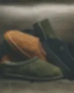 An artistic still-life photograph featuring a pile of UGG Tasman Woven slippers arranged on a textured grey leather surface. The image displays the three available colorways: two olive green slippers (Forest Night) in the foreground and top, one classic tan slipper (Chestnut) in the center-left, and a black slipper (Black) on the right. Each slipper prominently showcases its distinctive basket-weave textured suede upper, braided stitching around the ankle collar, and the UGG logo embossed on the side