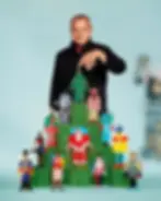 Studio portrait for Galeries Lafayette where French designer Jean-Charles de Castelbajac, dressed in a black jacket, stands behind a tiered green display featuring whimsical, paper-cutout holiday figures and a central Christmas tree, gently touching the top of the tree in his signature pop-art festive style
