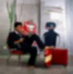 Portrait of artist Jean-Michel Basquiat barefoot and wearing a dark suit, sitting on a red chrome chair inside his art studio. His Neo-Expressionist style paintings can be seen in the background