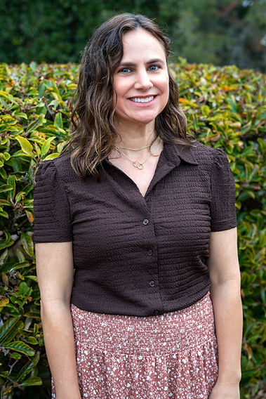 Portrait of Kathleen Jackson smiling, wearing a brown blouse and patterned skirt, standing outdoors by greenery.