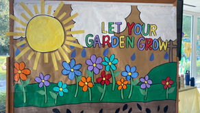 Colorful handmade “Let Your Garden Grow” display with a bright sun, clouds, raindrops, flowers, green hills, and seeds, created for Summer Arts Camp.