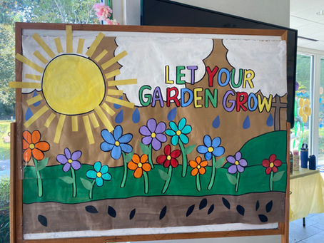 Colorful handmade “Let Your Garden Grow” display with a bright sun, clouds, raindrops, flowers, green hills, and seeds, created for Summer Arts Camp.