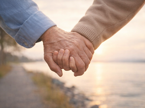 An older couple holding hands while walking beside calm water at sunset.