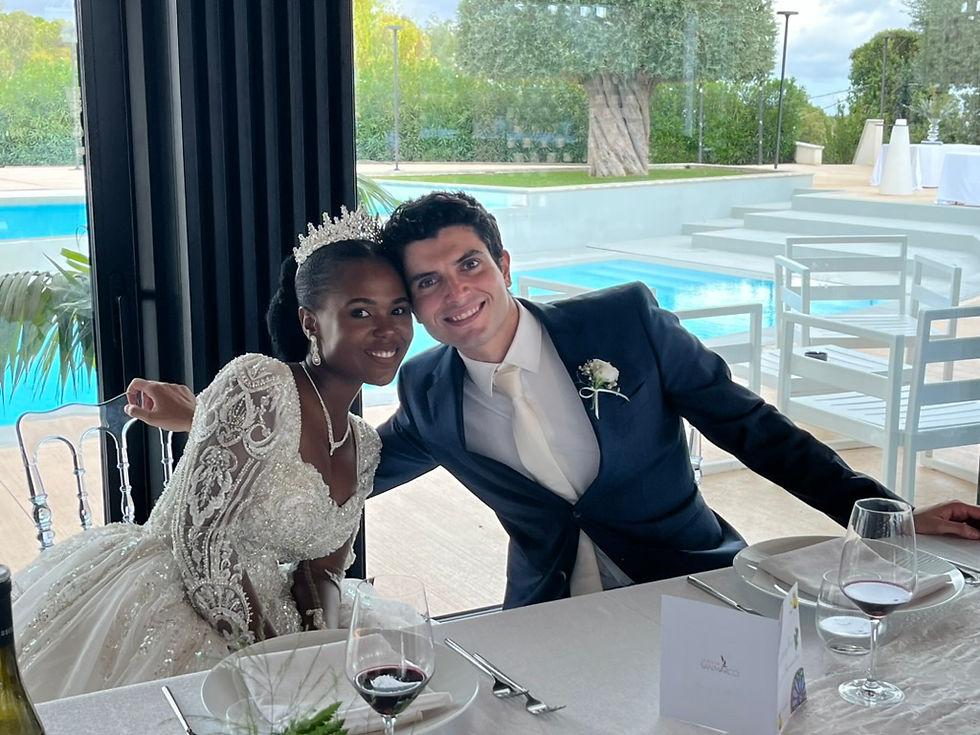 Bride in a white gown and groom in a blue suit smile at a table by a pool. Elegant setting with wine glasses and greenery outside.