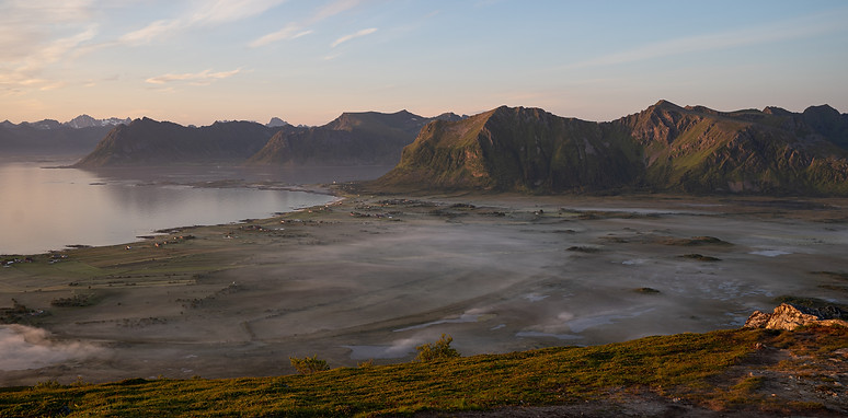 En spektakulær utsikt over et norsk fjordlandskap badet i mykt morgenlys. Dalen er dekket av lavtliggende tåke, omgitt av majestetiske fjell som speiler seg i stille vann. Bildet formidler en følelse av fred og naturens overveldende skjønnhet, perfekt for historier om utforskning og bærekraftig samspill med naturen. Utforsk flere unike perspektiver med [Tokt Film]. Hoven, Lofoten