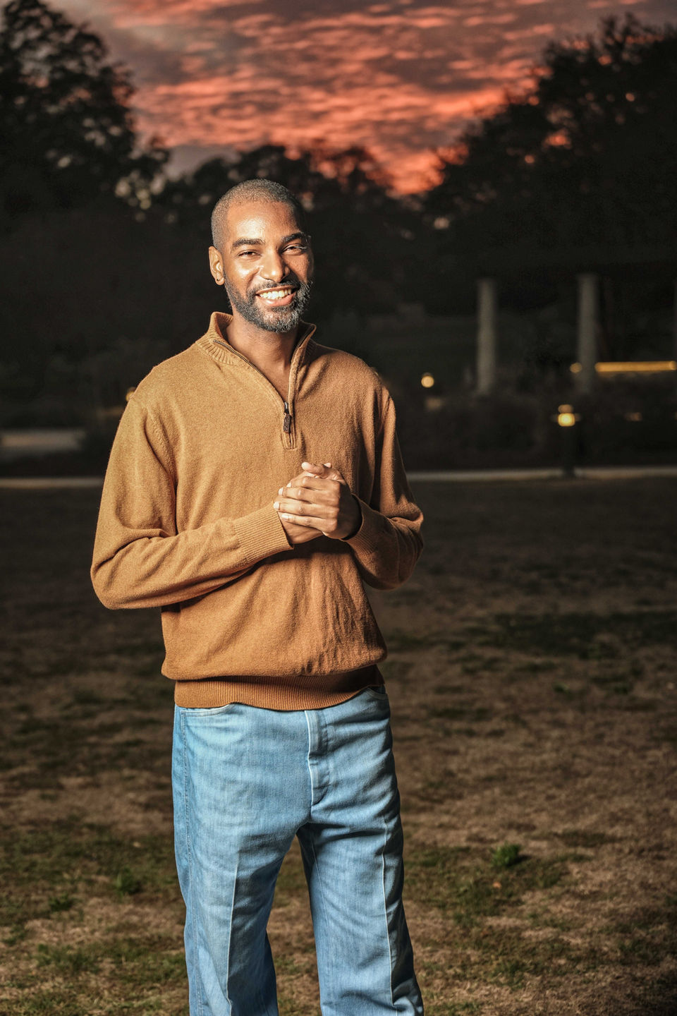 a portrait of a man in a long sleeve shirt and jeans during golden hour, the background is illuminated with red and orange hues of the setting sun.