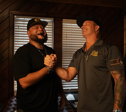 Scott Mason and Sergio Melendez smiling, wearing matching Jefe Cigar Lounge logo caps and shirts, holding cigars.