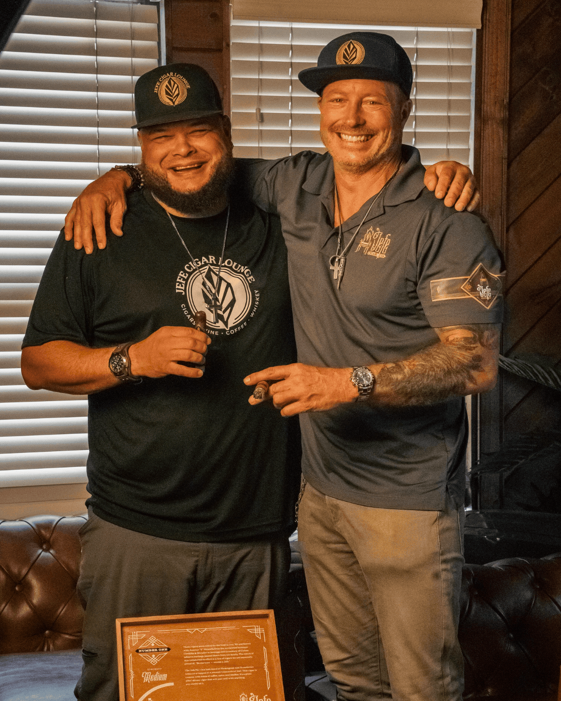 Scott Mason and Sergio Melendez smiling, wearing matching Jefe Cigar Lounge logo caps and shirts, holding cigars.
