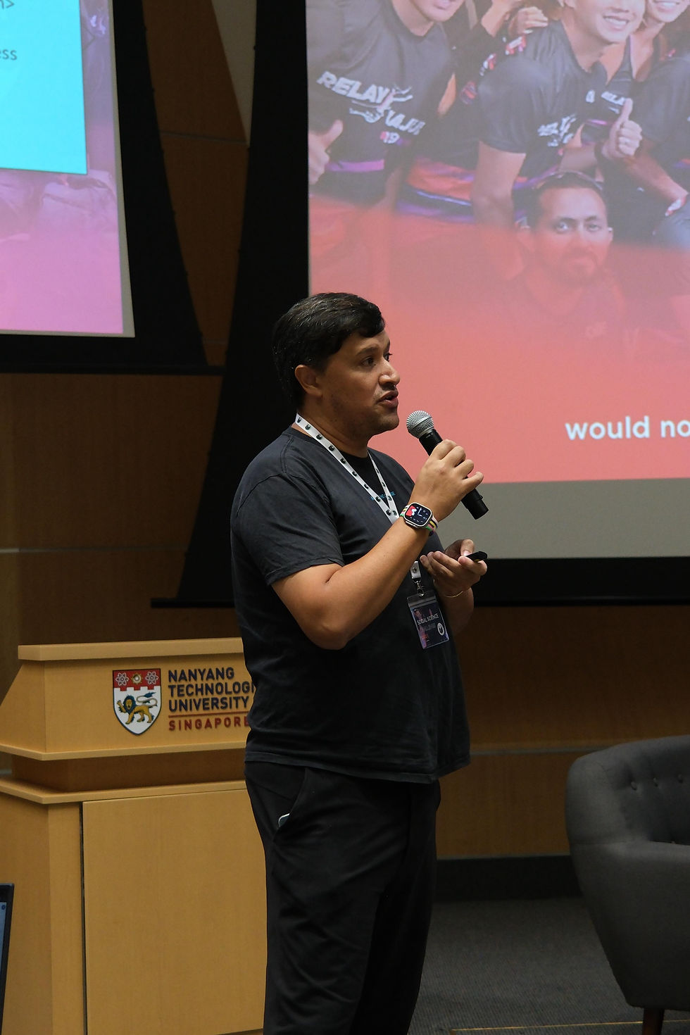 Man speaking into a microphone in a lecture hall at Nanyang Technological University, Singapore. Red and blue slides visible in the background.