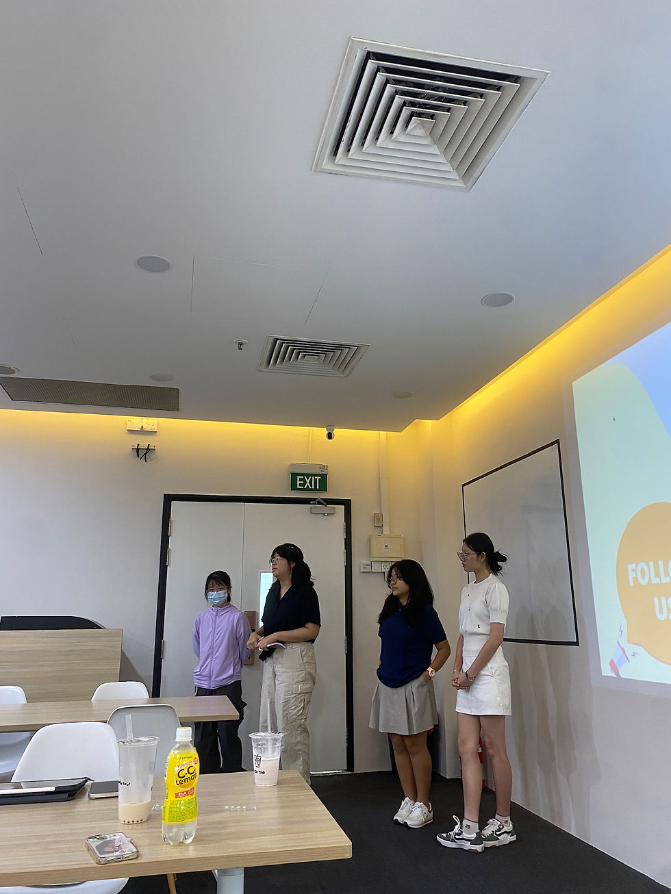 Four people standing in a classroom, presenting by a screen with "FOLLOW US" text. Bright-lit room with exit sign visible. Casual attire.