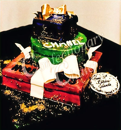 Three-tiered cake decorated as gift boxes with a Chanel theme, in blue, green, and red with white ribbons, set against a dark backdrop.