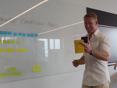Man smiling in an office, holding cards, standing by a whiteboard with blue and yellow sticky notes. Blurred text in the background.