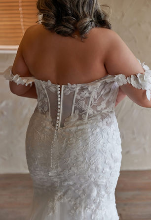 "Step into your fairytale with this enchanting plus-size wedding gown. The off-the-shoulder neckline, adorned with delicate floral lace, gently frames the shoulders with romantic softness. A sheer, intricately detailed bodice with supportive boning enhances and flatters your curves, while the mermaid silhouette and sweeping chapel train, embellished with 3D floral appliqués, bring both drama and romance. A true celebration of your beauty, this gown turns every step down the aisle into a magical moment."