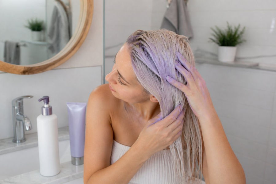 Woman applying purple shampoo to her icy blonde hair in a bright bathroom with conditioner on the counter