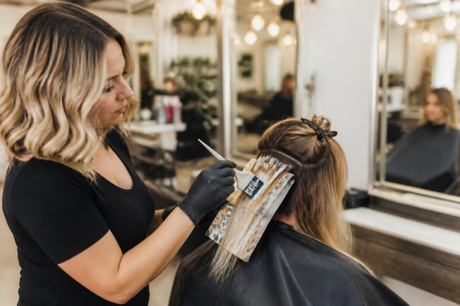 A professional colorist applying toner to a client's hair in a bright, modern salon, with mirrors and styling stations in the background