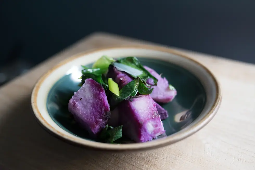 Purple yam pieces with green garnish on a dark ceramic plate, set on a wooden table. The mood is rustic and appetizing.