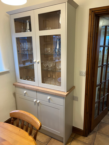 Light grey kitchen display cabinet, glass doors, wooden top, filled with glassware.