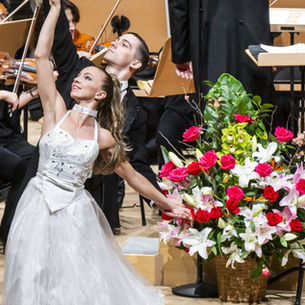 A ballet dancer in white stands poised next to an arrangement of pink and white flowers.