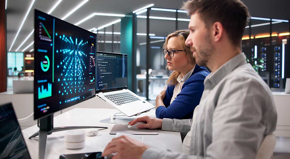 Two coworkers analyze data and code on dual monitors in a sleek office, with blue charts glowing on screen.