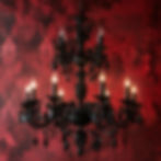 A black ornate chandelier in front of a dark red damask wallpapered wall.