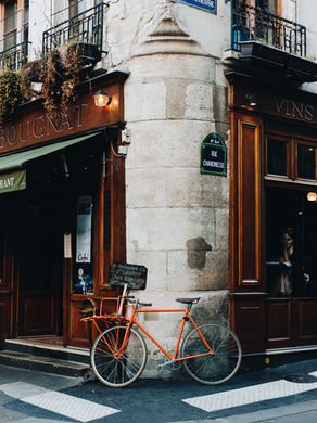 Orange bike leans against a Parisian café with wooden facade. Signs read "Rue Chanoinesse" and "Restaurant Inside." A person peers out.