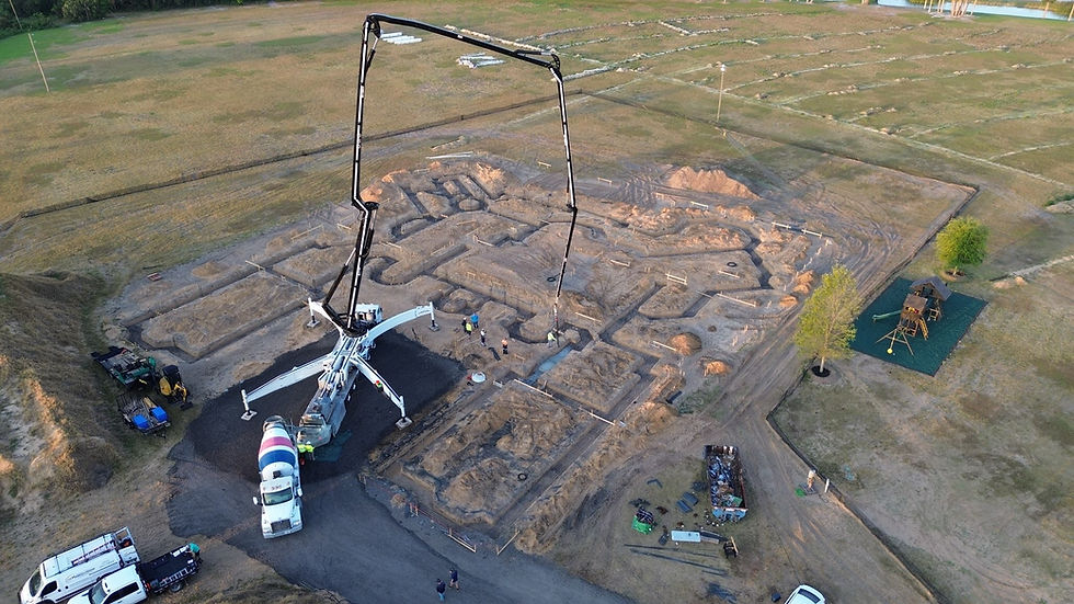 Aerial view of construction site with a large concrete pump and mixer, workers in safety gear, surrounded by grassy fields and trees.