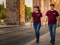 Dos brigadistas de Morena caminando por la banqueta junto a los Arcos de Querétaro al atardecer, representando el desafío electoral del partido en la entidad.