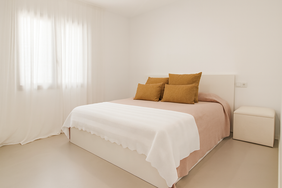 Minimalist bedroom with blush bedspread, mustard accent pillows, white throw blanket, and sheer curtains allowing natural light – Can Ferragut Mallorca.