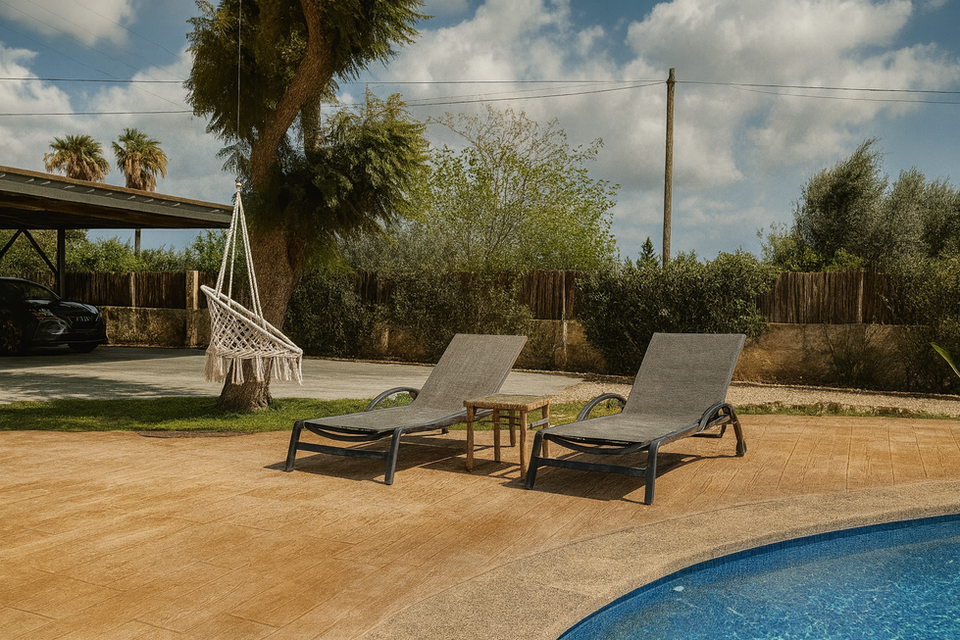 Poolside relaxation area with two gray loungers, small wooden table, and hanging hammock chair under a tree – Can Ferragut Mallorca.