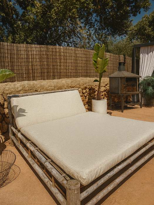 Outdoor bamboo daybed with white cushion, potted plants, rustic stone wall, and shaded pergola lounge in the background – Can Ferragut Mallorca.