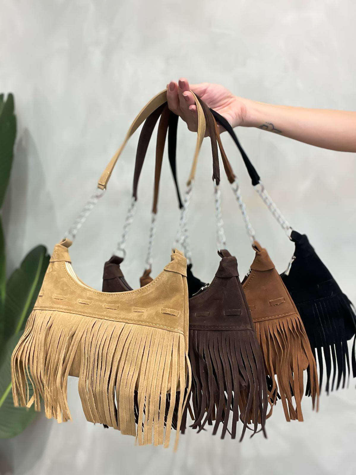 Mão segura cinco bolsas de ombro meia-lua com franjas em camurça nas cores: bege, marrom-escuro, marrom-médio, caramelo e pre