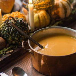 A pot of creamy orange soup with a ladle surrounded by pumpkins, gourds, and lit candles on a wooden table, creating a cozy autumn vibe.